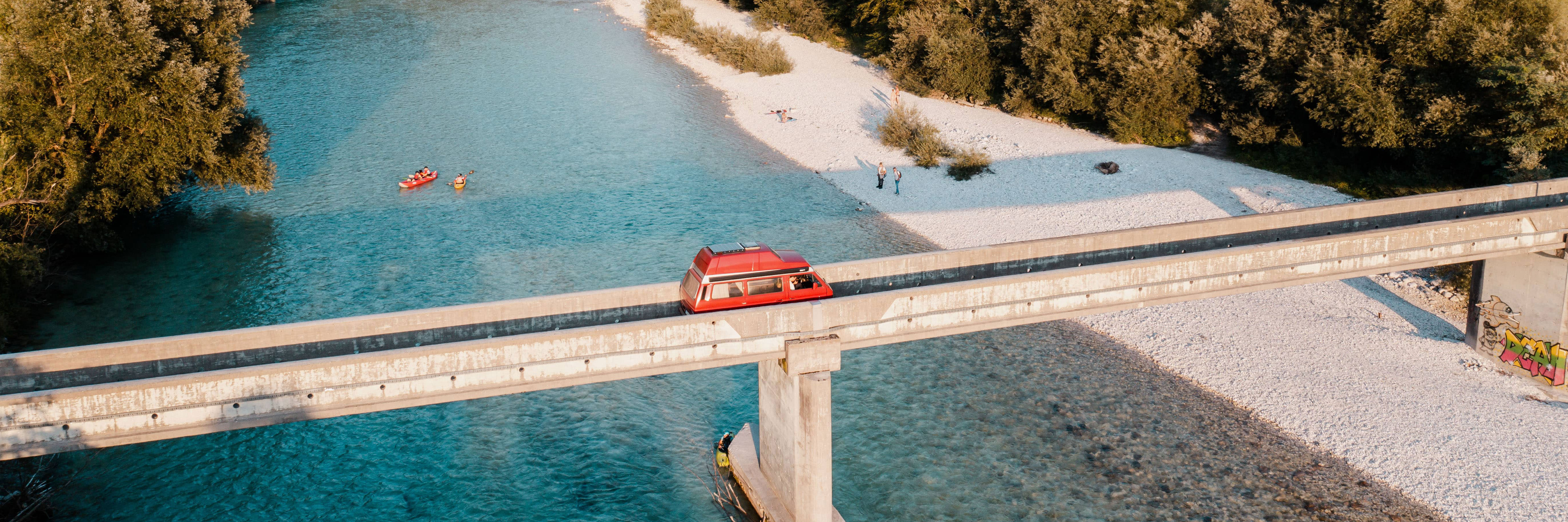 Ein rotes Wohnmobil fährt in der Abend-Sommersonne über eine schmale Brücke über einem glasklaren Fluss